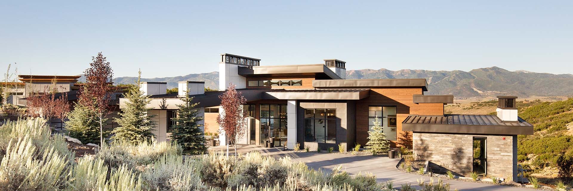 Contemporary luxury mountain home on a hillside with wood-and-stone exterior, large windows, evergreen landscaping and panoramic mountain views.