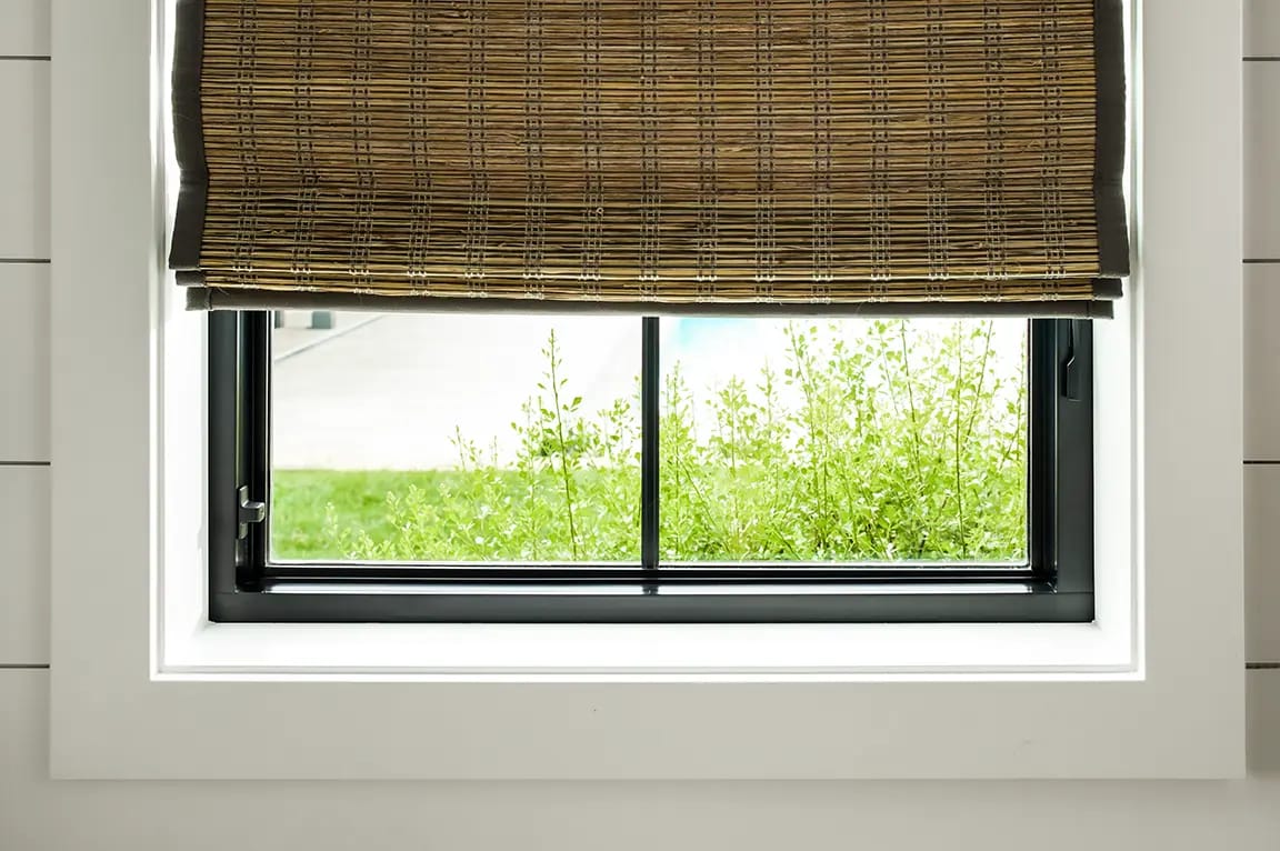 Black-framed window with white trim and a partially lowered bamboo Roman shade overlooking green shrubs