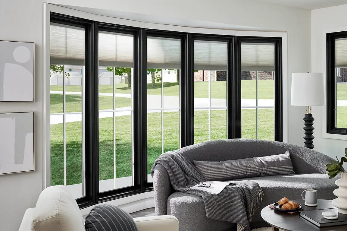 A modern living room featuring a black bow window, a curved gray sofa and a view of the front yard.