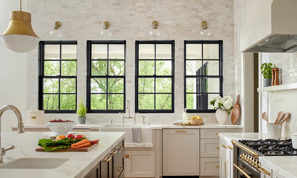 A modern kitchen with stone-accent walls with three black casement windows showing trees outside.