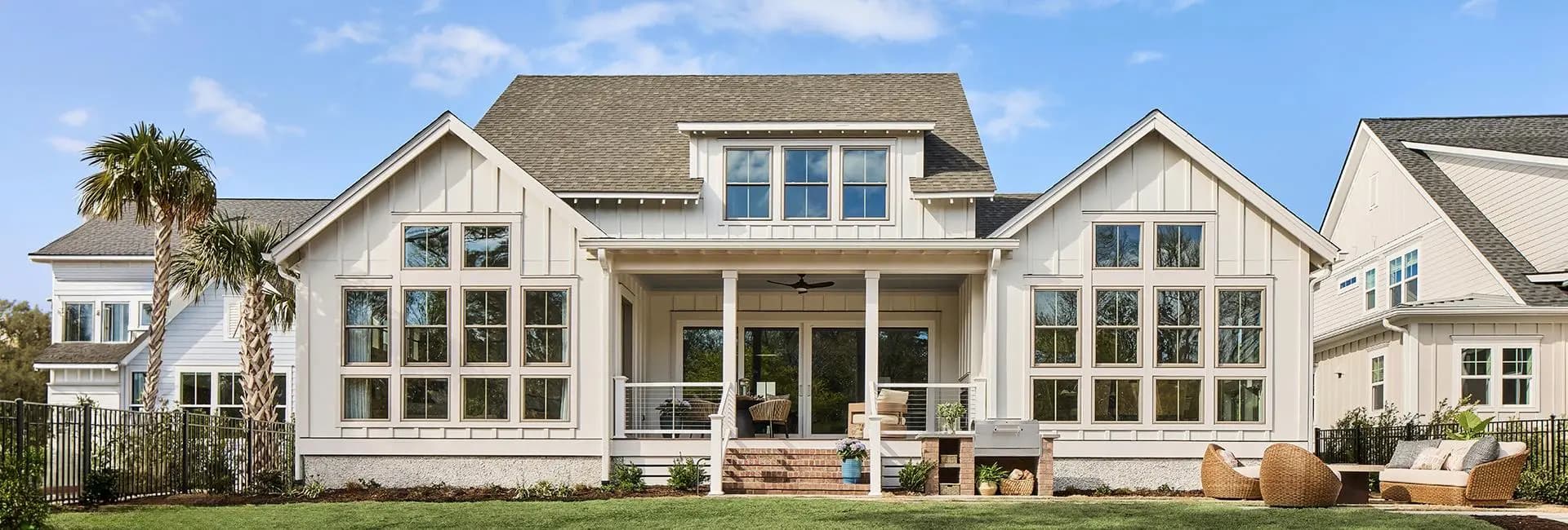 Modern coastal farmhouse exterior with white board-and-batten siding, large grid windows, covered porch, palm trees and outdoor seating on a landscaped lawn.