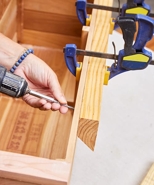 Two clamps are holding a board in place while a long screw is being fastened to securely attach to the window box.
