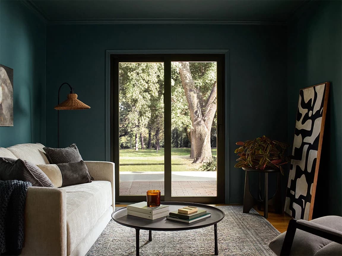 A modern living room with a large sliding door opening to a green yard, featuring a sofa, coffee table, and abstract art.