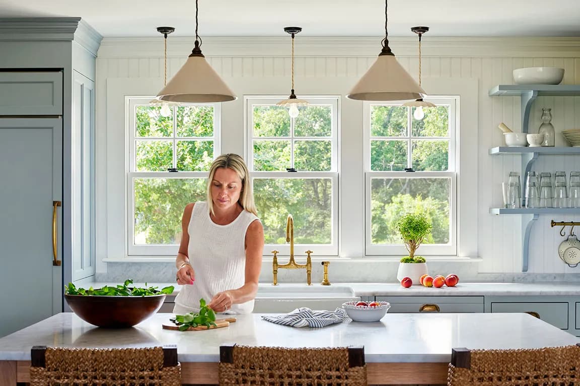 Bright kitchen with white wood windows, gold faucet, and a woman preparing food at a marble island.