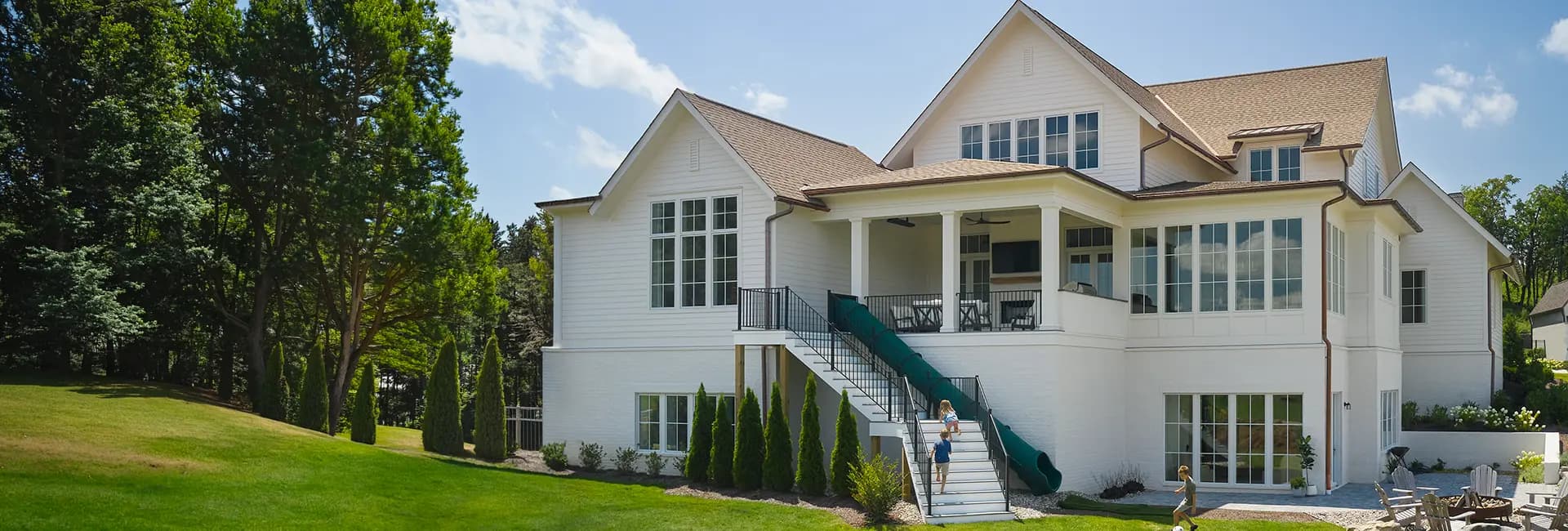White two-story modern farmhouse with covered porch, exterior staircase featuring a green slide, and children playing on the landscaped backyard lawn.