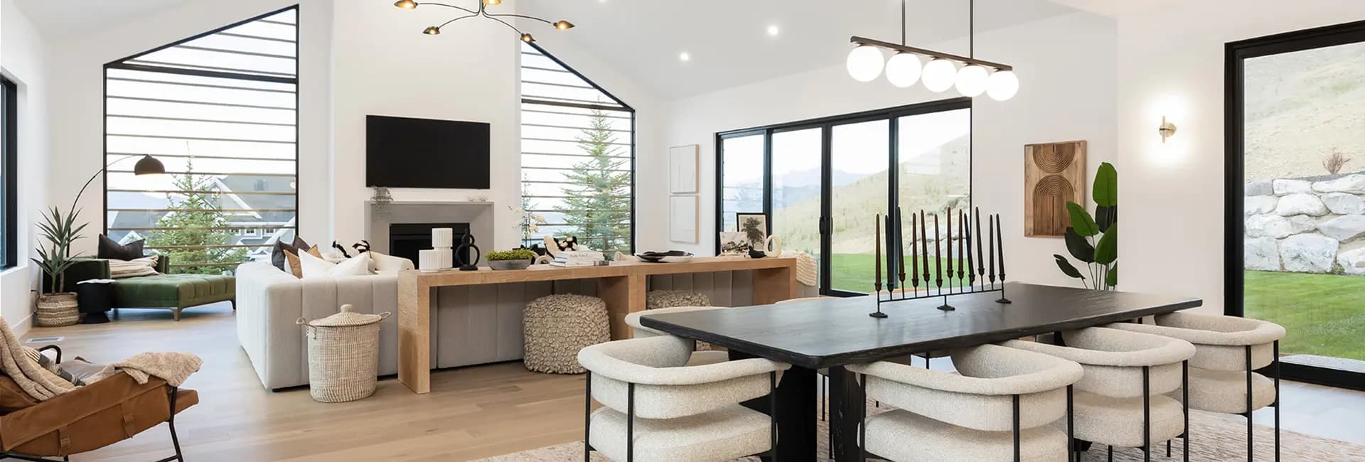 Modern open-concept living/dining room with black table and cream chairs, white sectional, fireplace, and large windows with hillside view