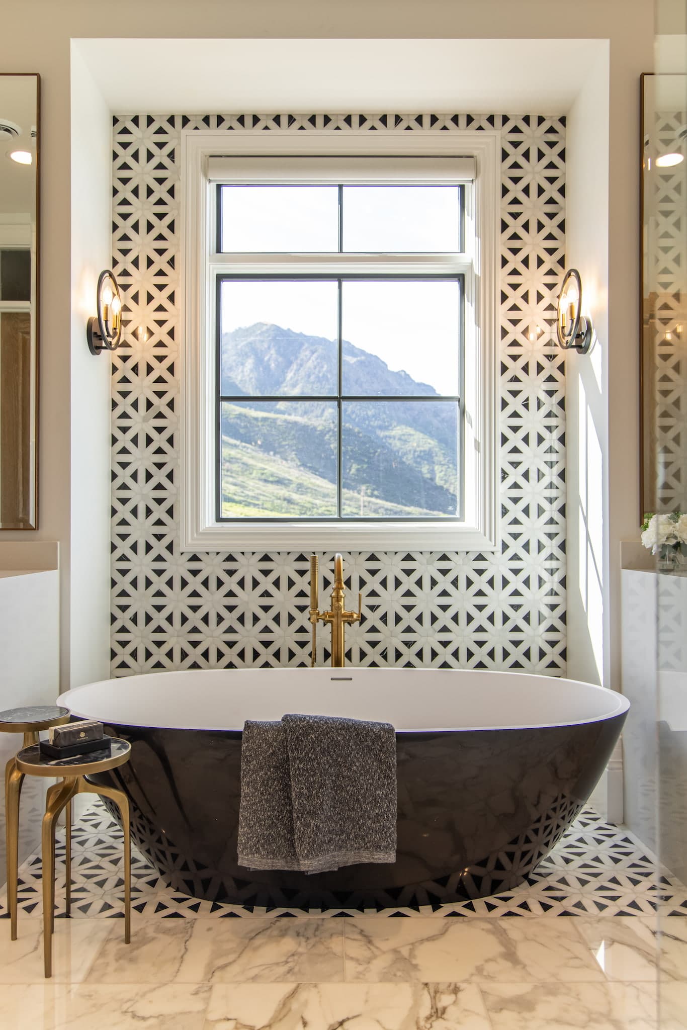 This freestanding bathtub with gold hardware sits on patterned tile with a picture window above it.