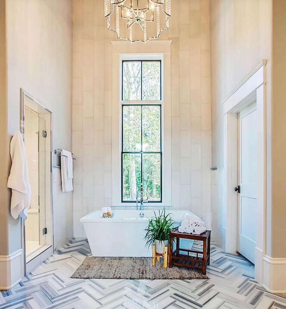 A modern and contemporary bathroom with high ceilings and a chandelier features a bathtub with a tall black window above.