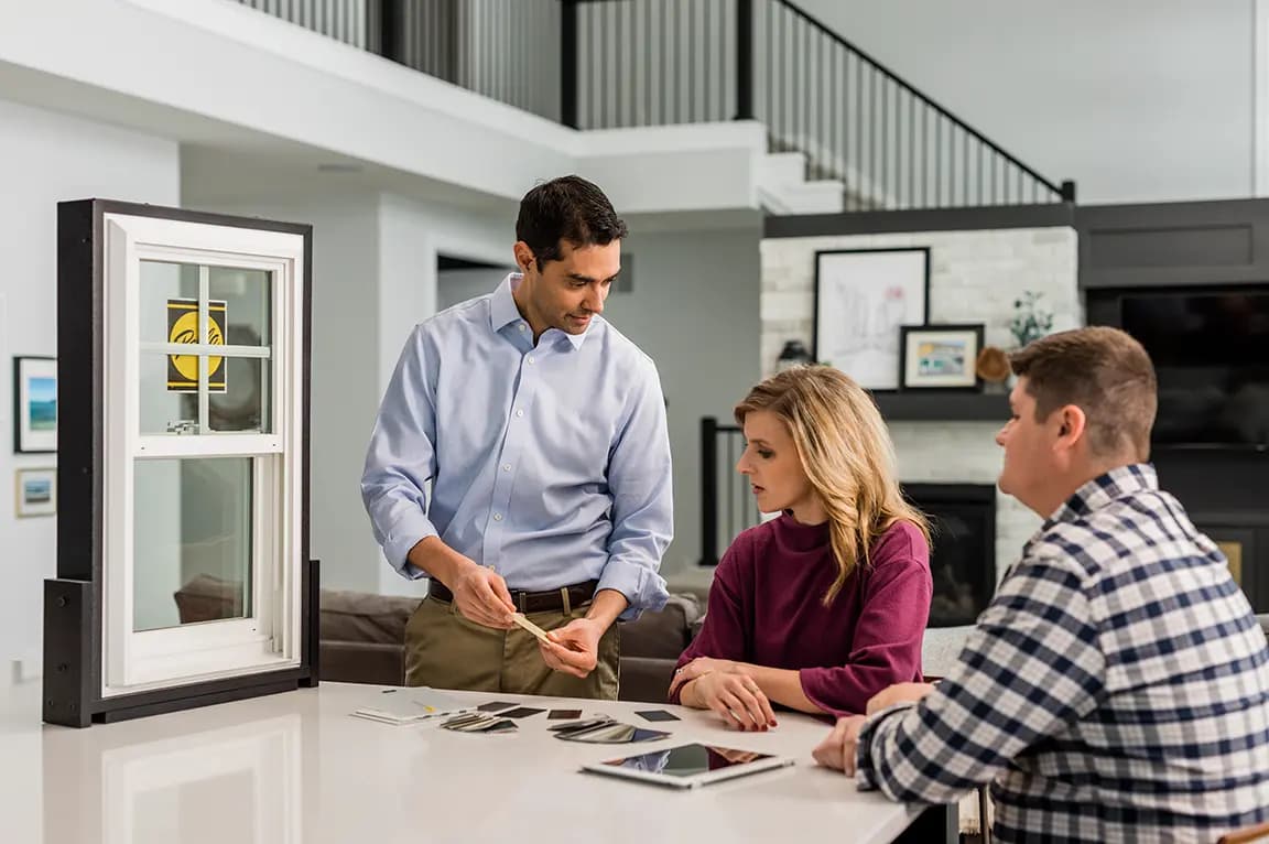 Window replacement consultant showing vinyl window samples and color swatches to homeowners at a kitchen island in a modern open-plan home.