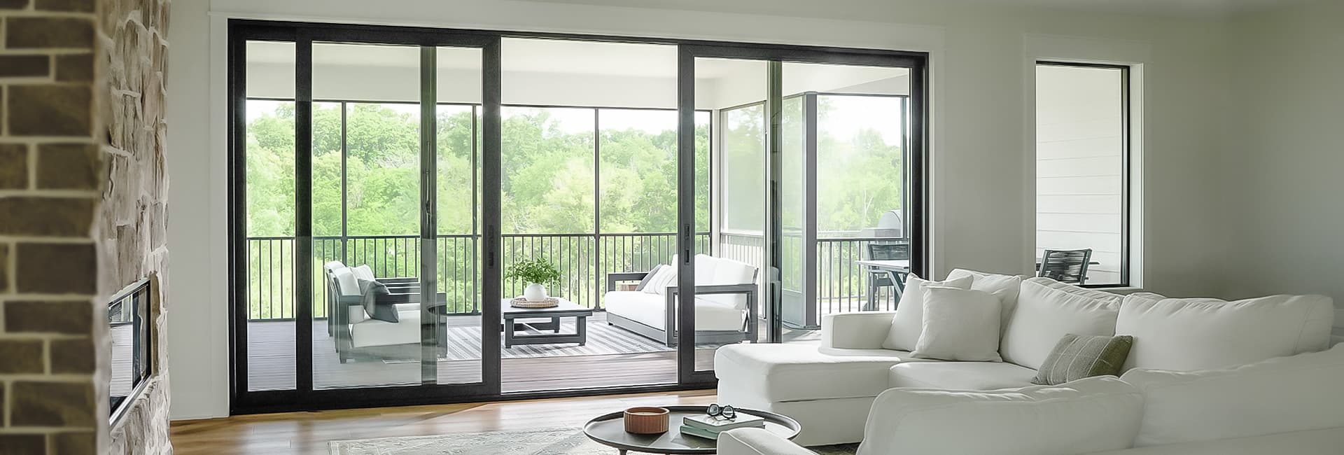 Modern living room with white sectional sofa and black-framed sliding glass doors opening to a covered patio with outdoor seating and a wooded view.