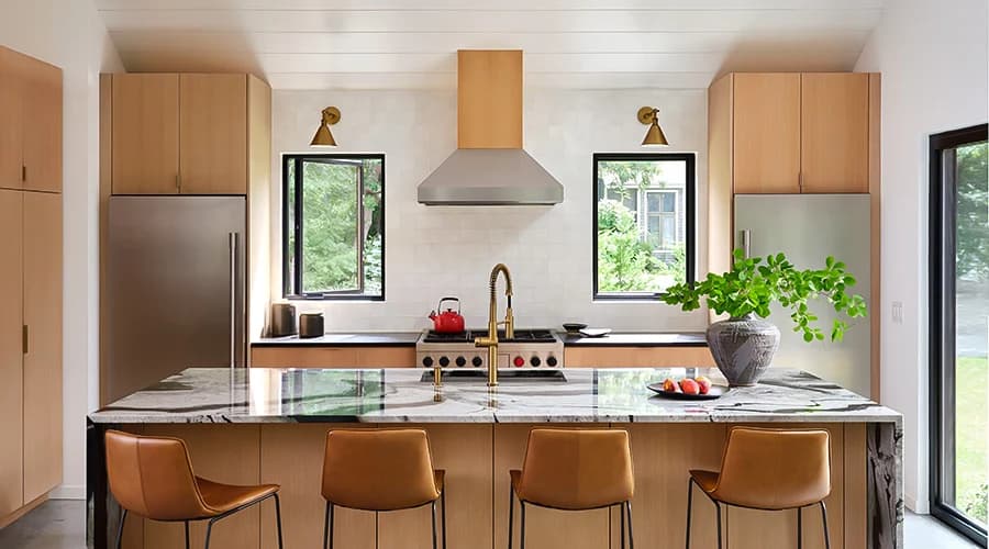 A modern kitchen with wood cabinets, marble island, and bright weather visible through two windows.