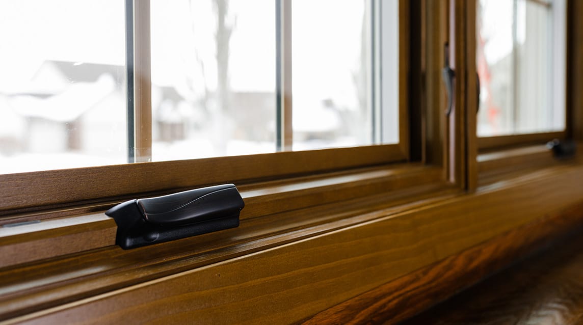A hand holding a paintbrush staining the bottom sash of a wood double-hung window.