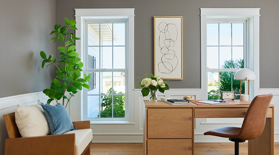 A modern office featuring large windows, a wooden desk, a plant, and decorative art, showcasing natural light.