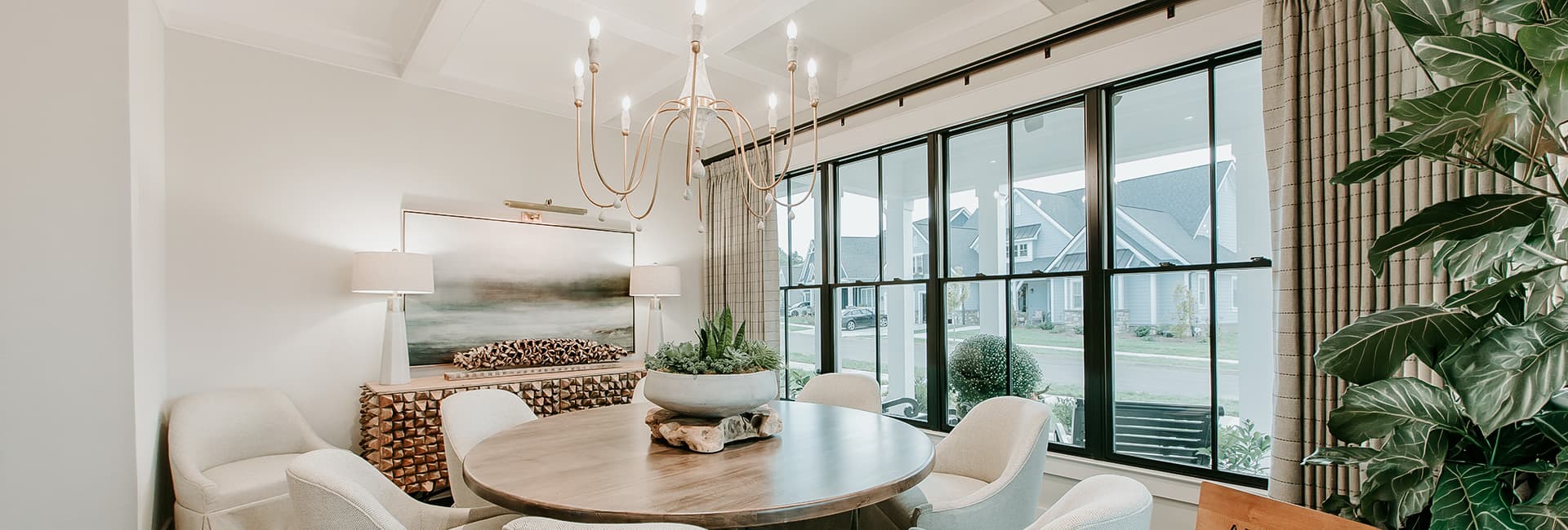 Modern dining room with round wooden table, upholstered chairs, brass chandelier, large black-framed windows and indoor plants.