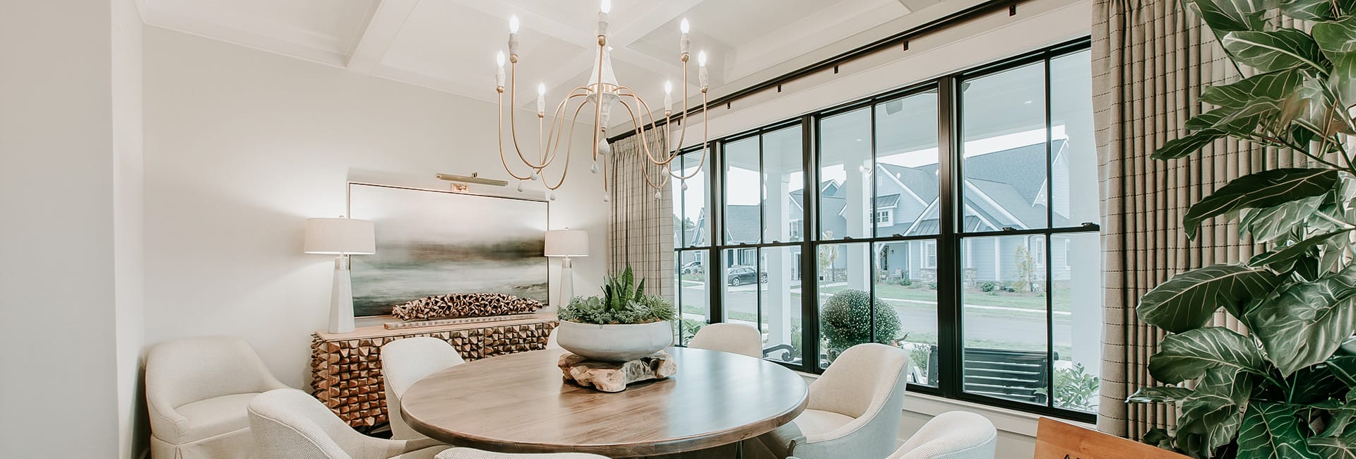 Modern dining room with round wooden table, upholstered chairs, brass chandelier, large black-framed windows and indoor plants.