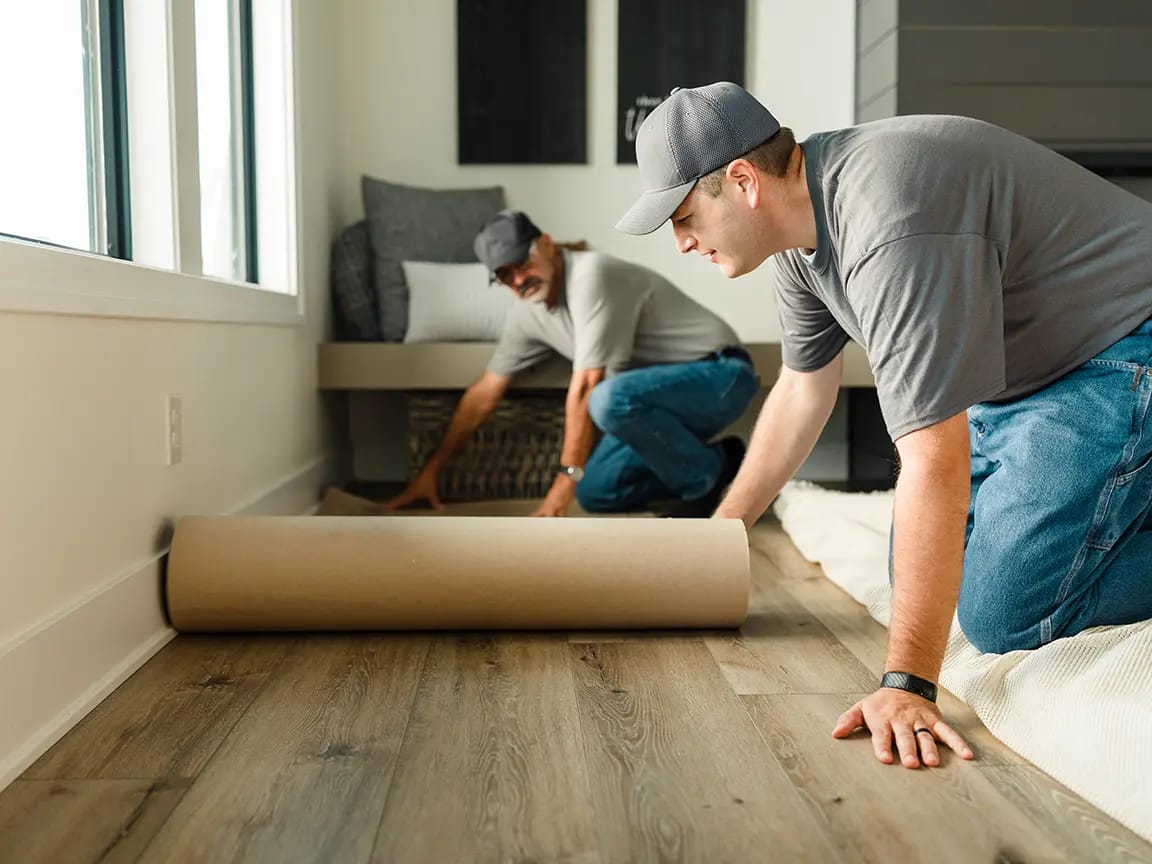 Two flooring installers installing wood-look vinyl plank flooring in a living room during a home renovation.