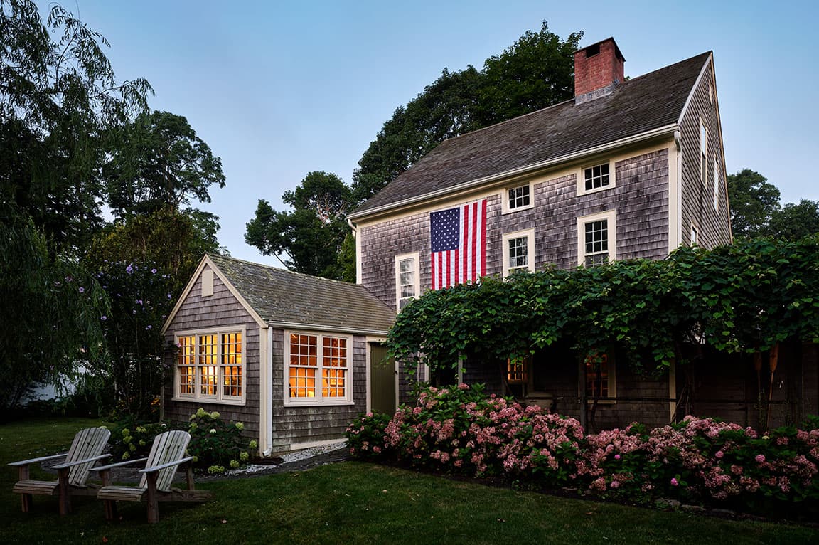 A charming cape cod home with an American flag and surrounded by lush greenery.