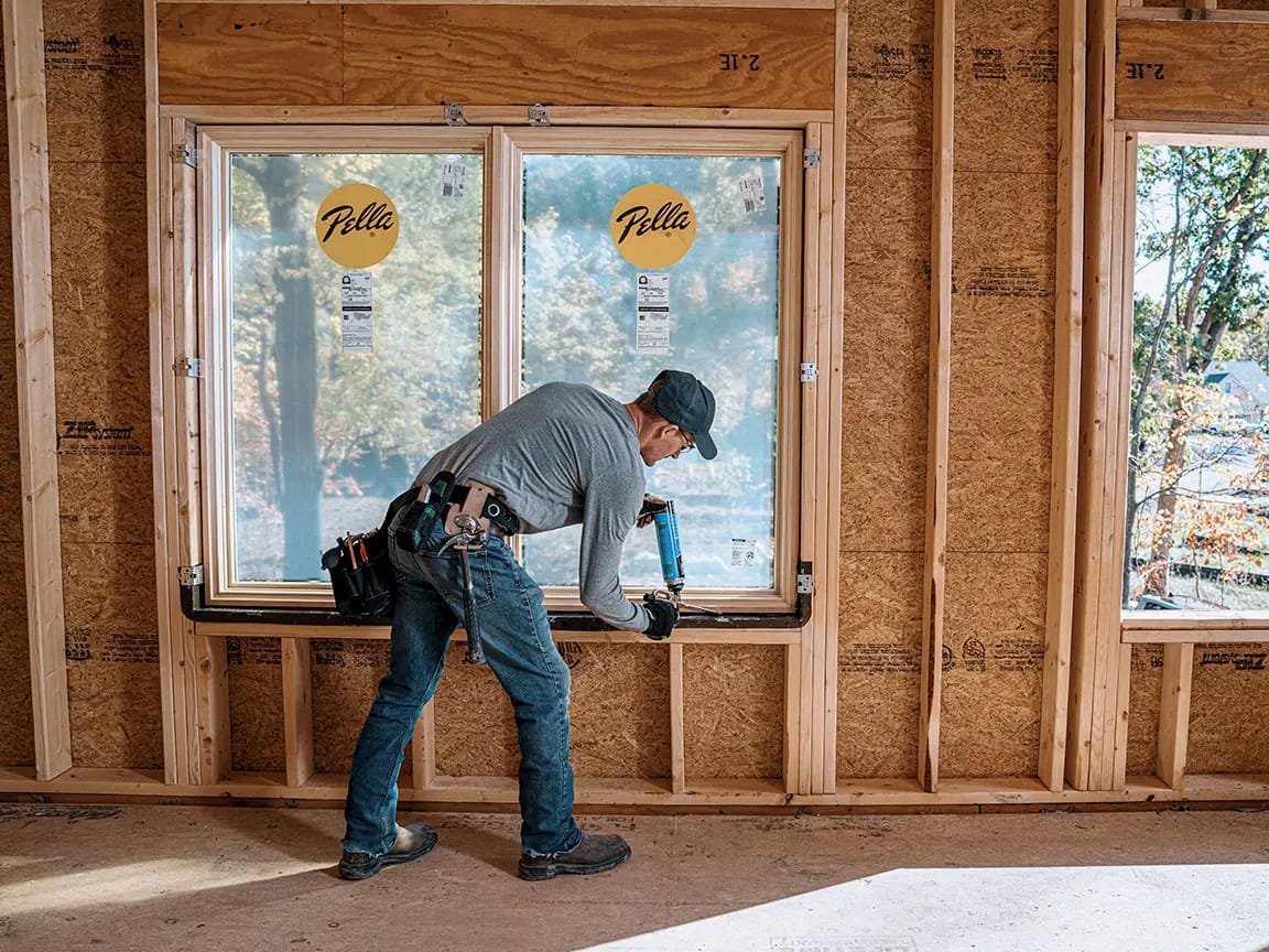 Construction worker applying sealant with a caulking gun to a newly installed Pella window in a wood-framed house under construction.