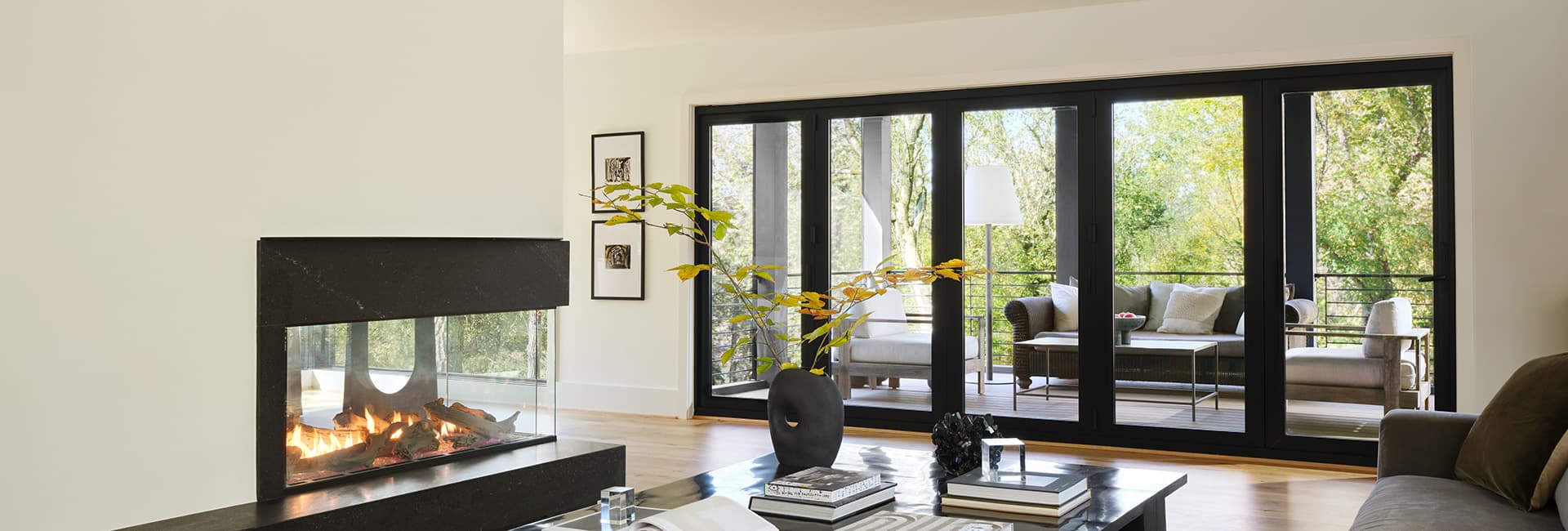 Modern living room with glass-front fireplace and black-framed folding doors opening to a tree-lined patio with seating.