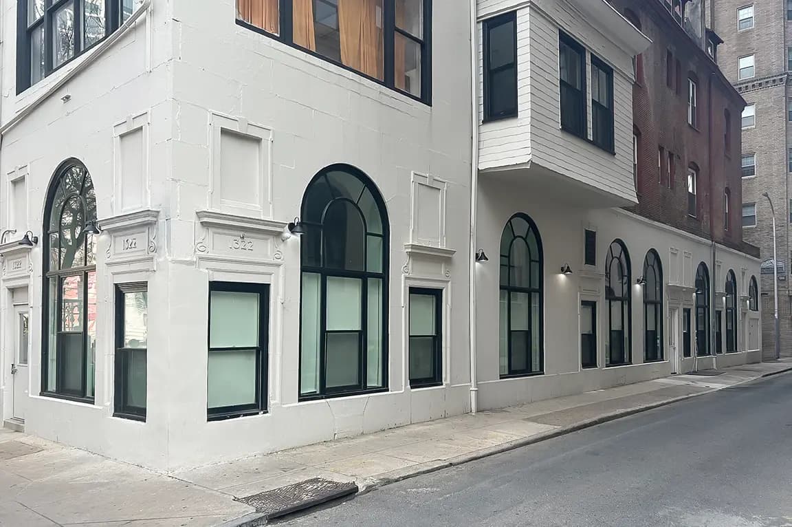 White corner building with tall arched black-framed windows and sidewalk on a quiet urban street