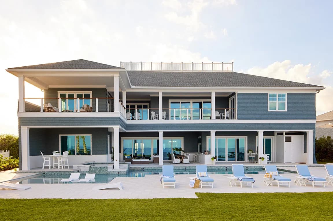 A modern coastal home with large hurricane doors, blue siding, and a poolside patio with lounge chairs.