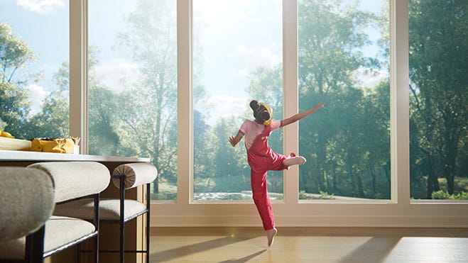 a girl wearing pink overalls is dancing in front of large windows while it is hot outside.