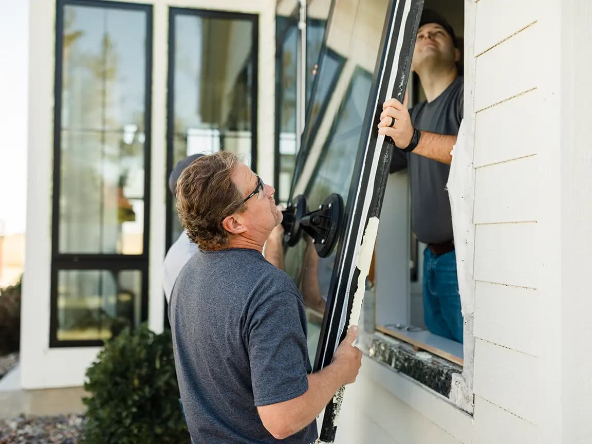 Window installation: two workers lifting a large glass pane with a suction cup lifter into a house window opening.