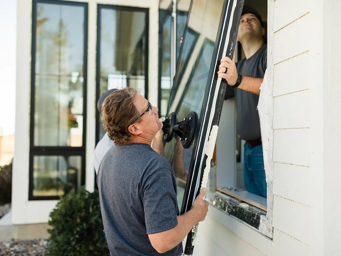 Window installation: two workers lifting a large glass pane with a suction cup lifter into a house window opening.