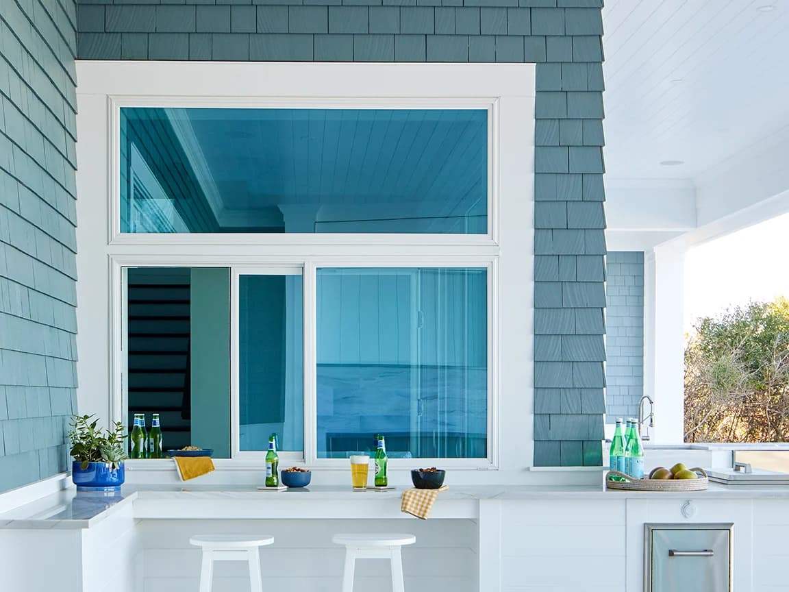 A hurricane impact window set in blue shingles above an outdoor bar with drinks and snacks.