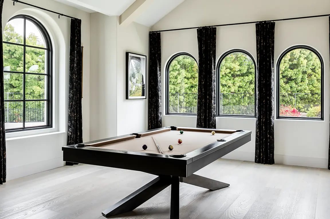 Modern game room with beige-felt pool table and black frame, light hardwood floors, black curtains and black-framed arched windows overlooking greenery.