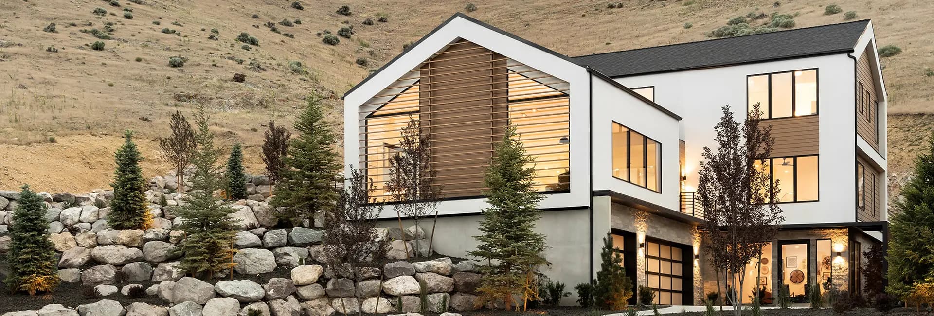 Modern three-story hillside home with large windows, wood-louvered facade, rock retaining wall and evergreen landscaping lit at dusk