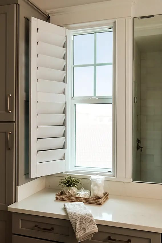 Bathroom vanity with white plantation shutters on window, gray cabinets, white countertop with wicker tray, potted plant, glass cotton jar and folded towel.