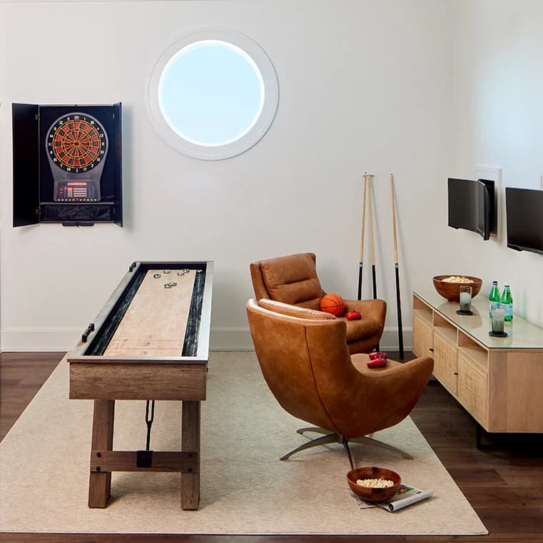 A modern game room with a dartboard, shuffleboard table, leather chair, and round window, not related to a hurricane.