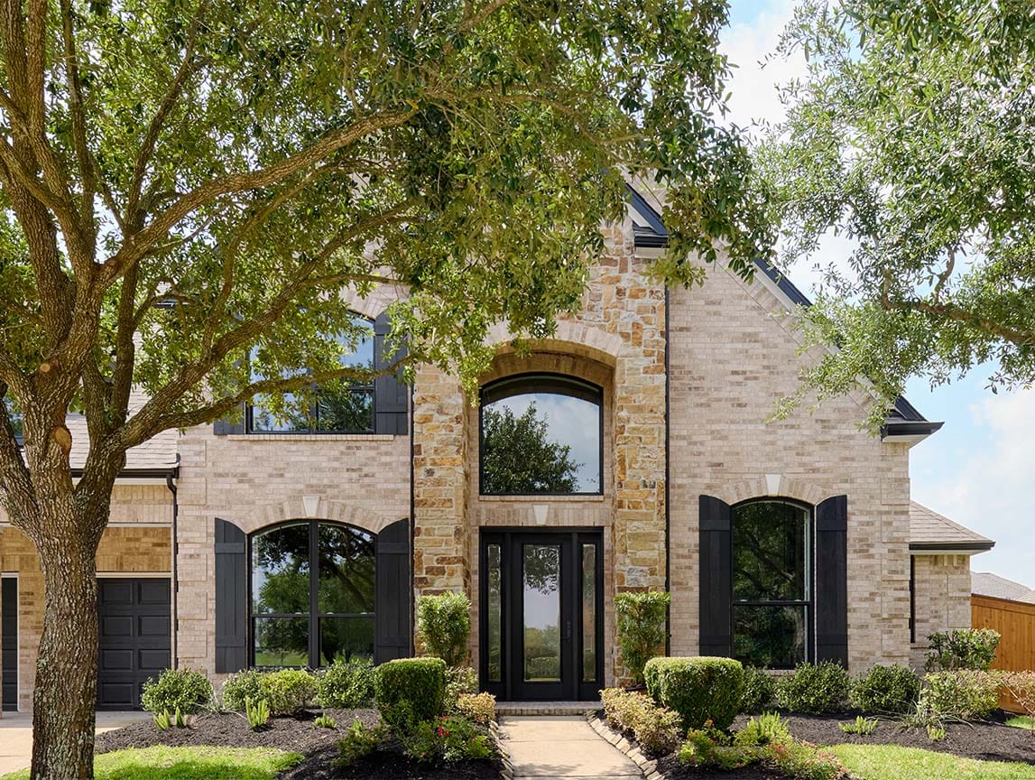 Stone exterior home with large black windows, arched entryway, and landscaped front yard framed by mature trees.