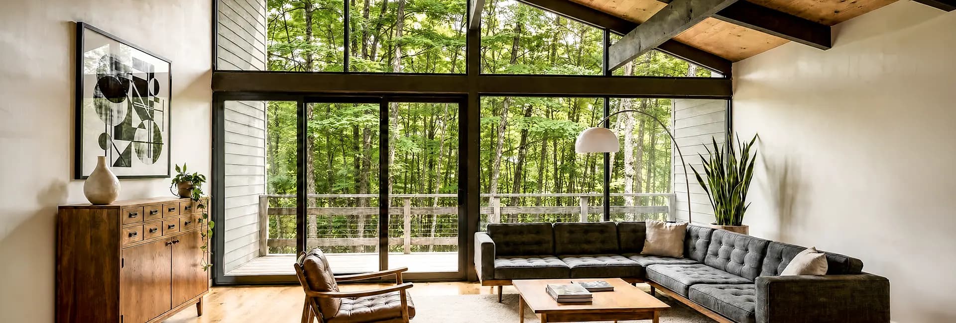 Modern living room with floor-to-ceiling windows overlooking a green forest, gray tufted sectional sofa, wooden coffee table and mid-century leather armchair