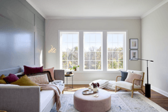 a living room with a dark accent wall on the left and a cream wall on the right has three white casement windows on the far side of the room.