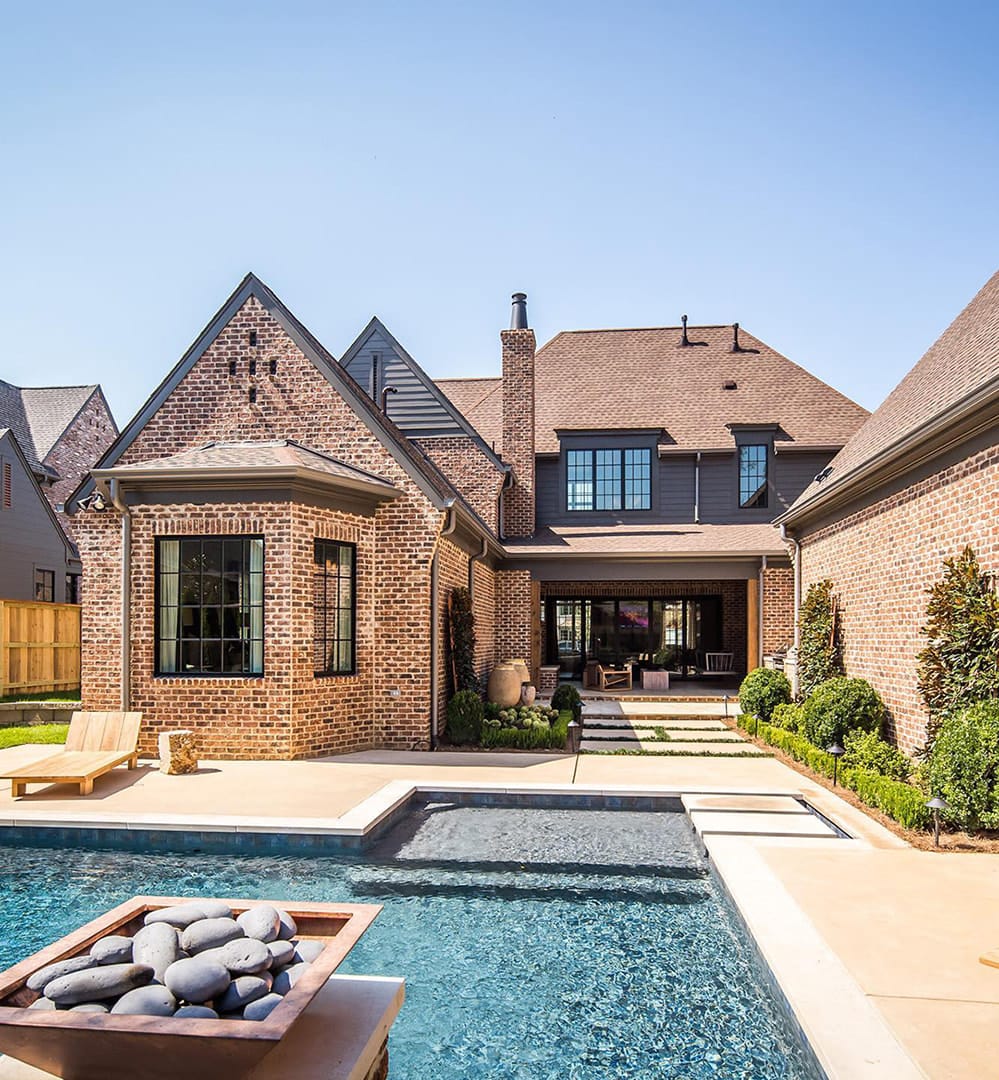 Beautiful pool and outdoor entertainment area in front of a red brick house with black windows.