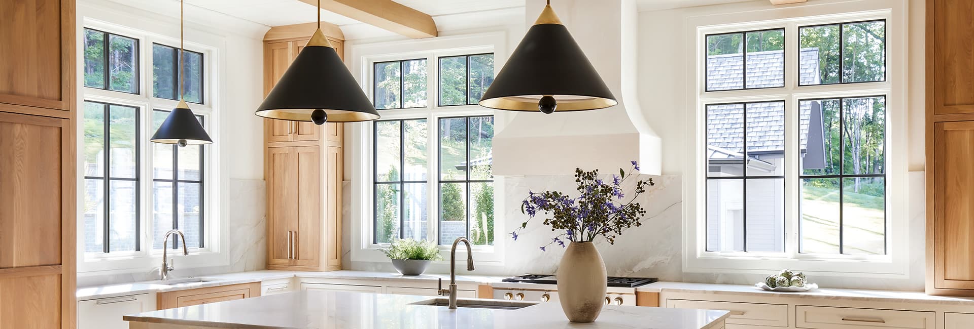Bright modern kitchen with natural wood cabinets, marble countertops, large black‑grille windows, black cone pendant lights, island sink with stainless faucet and vase of purple flowers