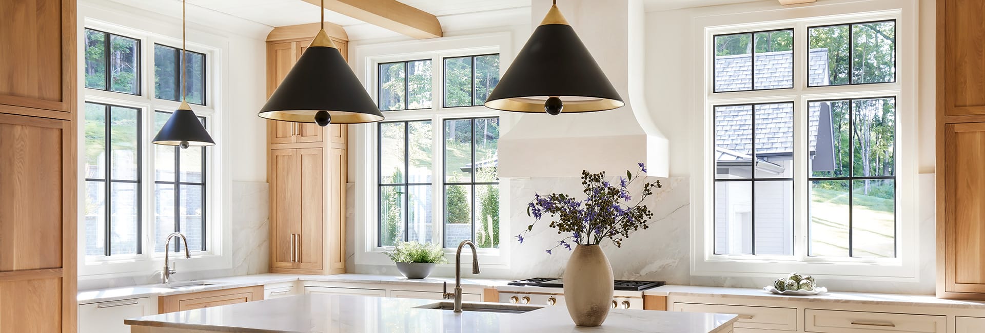 Bright modern kitchen with natural wood cabinets, marble countertops, large black‑grille windows, black cone pendant lights, island sink with stainless faucet and vase of purple flowers