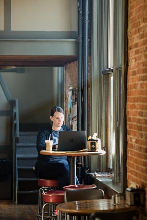 A woman is working at a tall coffee shop table, sitting next to tall windows.