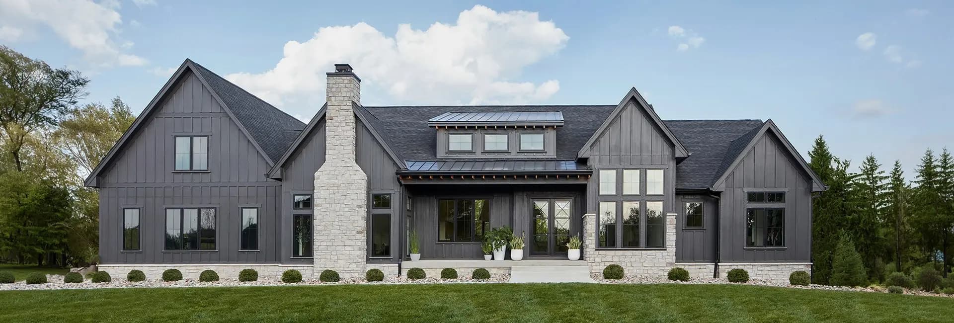 Modern gray farmhouse with board-and-batten siding, stone chimney, gabled roof, large windows and landscaped front lawn