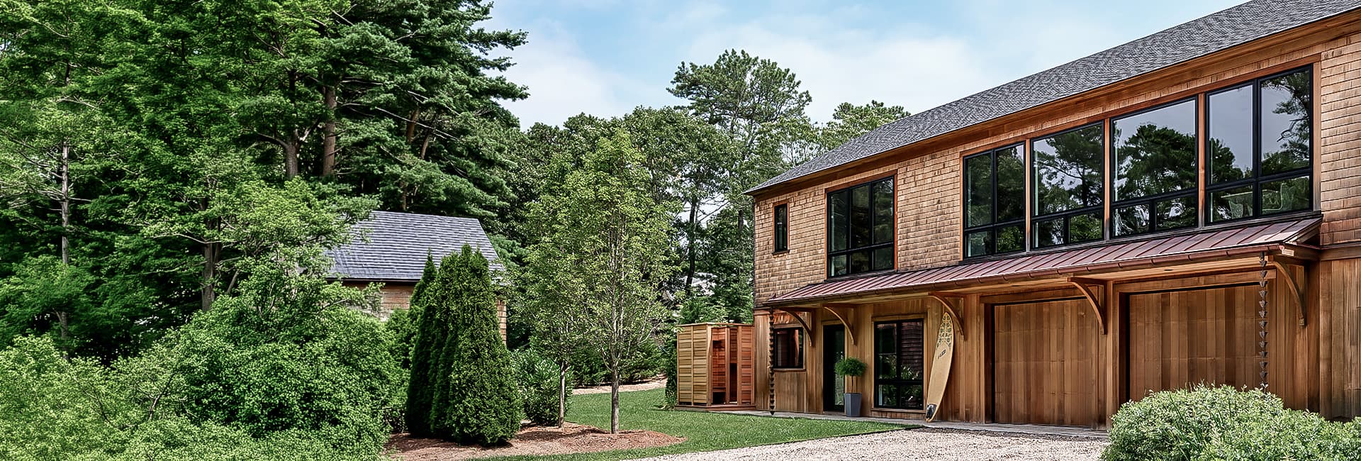 Modern cedar-clad two-story home with large windows and double garage, surfboard by entrance, landscaped yard and dense forest backdrop.