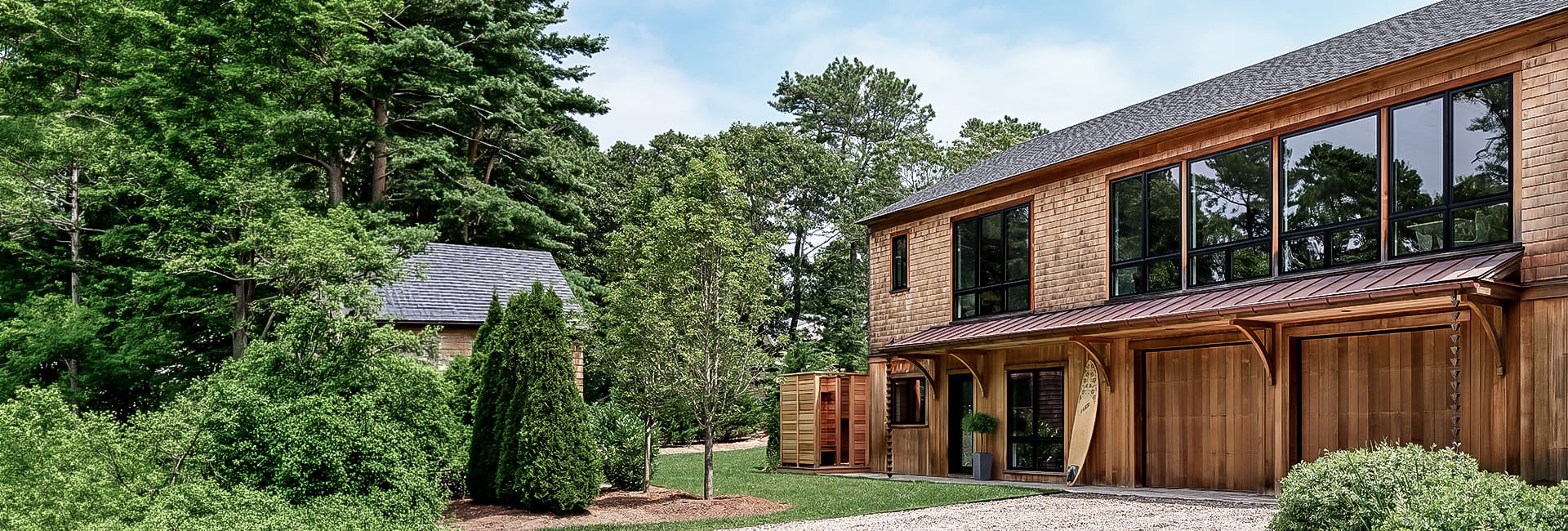 Modern cedar-clad two-story home with large windows and double garage, surfboard by entrance, landscaped yard and dense forest backdrop.