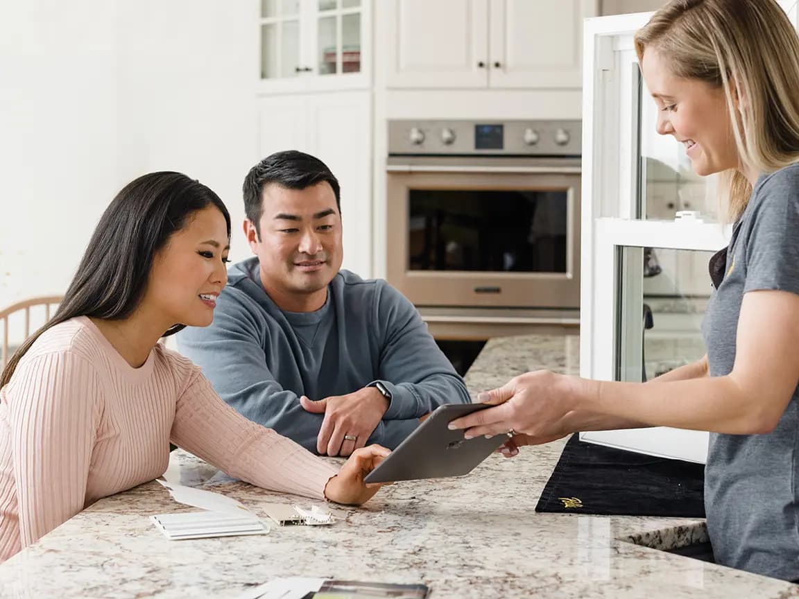 Couple at a bright kitchen island reviewing a tablet with a contractor during a home renovation consultation, samples on the countertop.