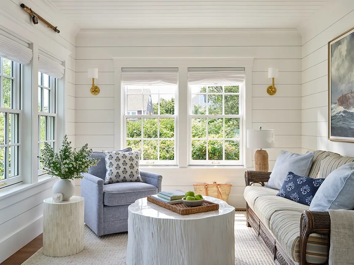 Sunlit coastal living room with white shiplap walls, large windows, blue armchair, striped sofa, round white textured coffee table and vase of greenery.