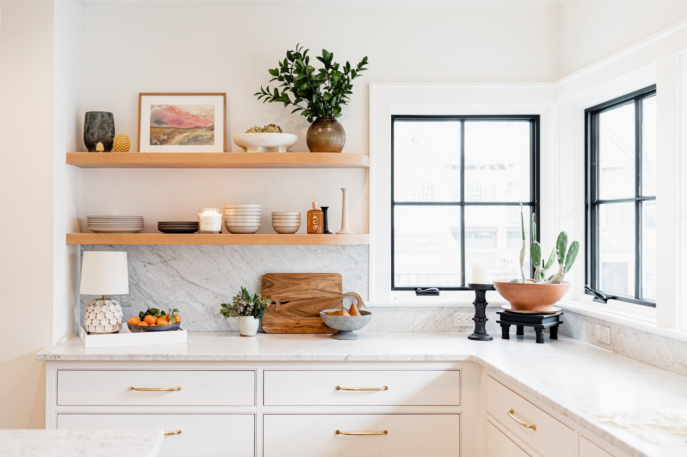 A white kitchen has open shelving next to two black square windows in the corner.