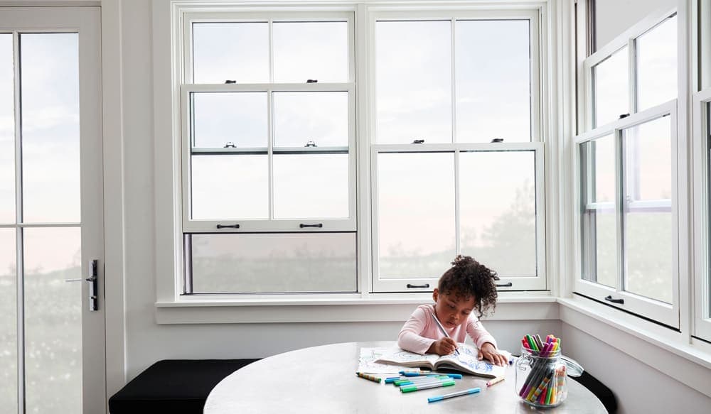 Pella's Integrated Rolscreen® Appears When You Need It girl coloring at table with white double-hung windows with integrated screen behind her