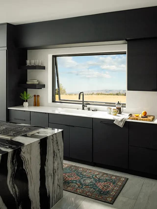 Modern black kitchen with marble island and gas cooktop, white countertop beneath a large casement window overlooking golden fields and distant hills.