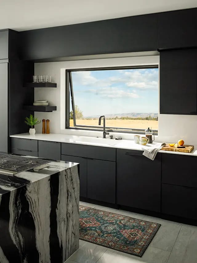 Modern black kitchen with marble island and gas cooktop, white countertop beneath a large casement window overlooking golden fields and distant hills.