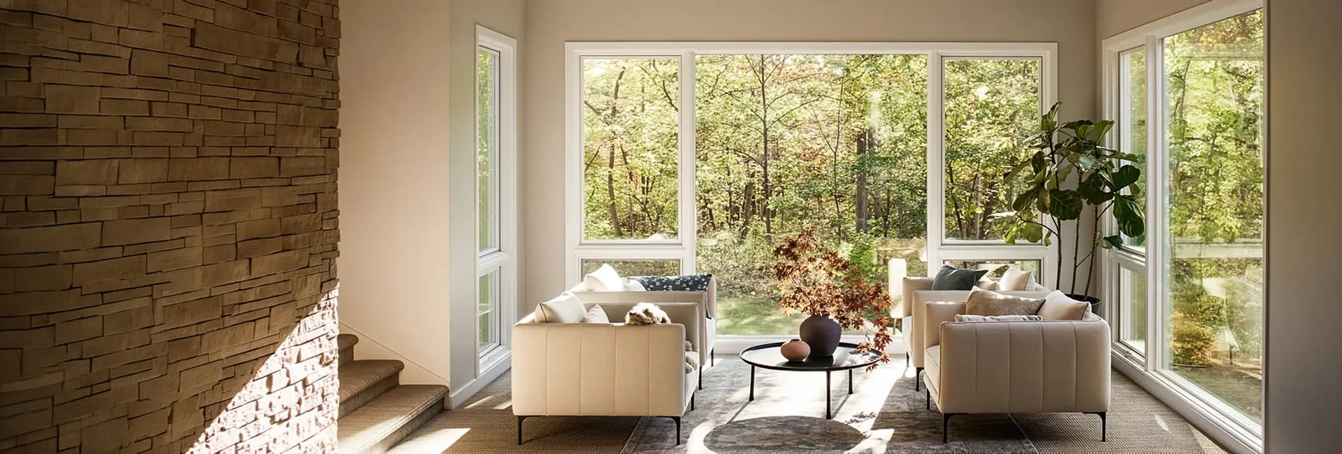 Modern living room with beige sofas, round coffee table and vase, floor-to-ceiling windows overlooking a forest.